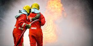 Two firefighters holding a fire hose in front of flames and smoke