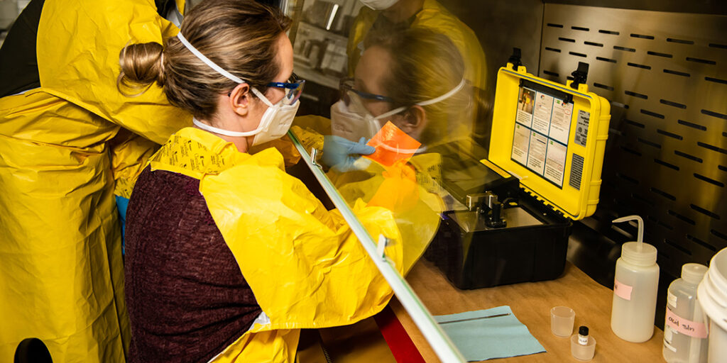 Two scientists in yellow PPE prepare samples under scientific hood